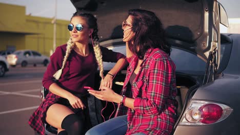 dos mujeres jóvenes atractivas con gafas de sol sentadas dentro del maletero del coche abierto en el estacionamiento junto al centro comercial y escuchando