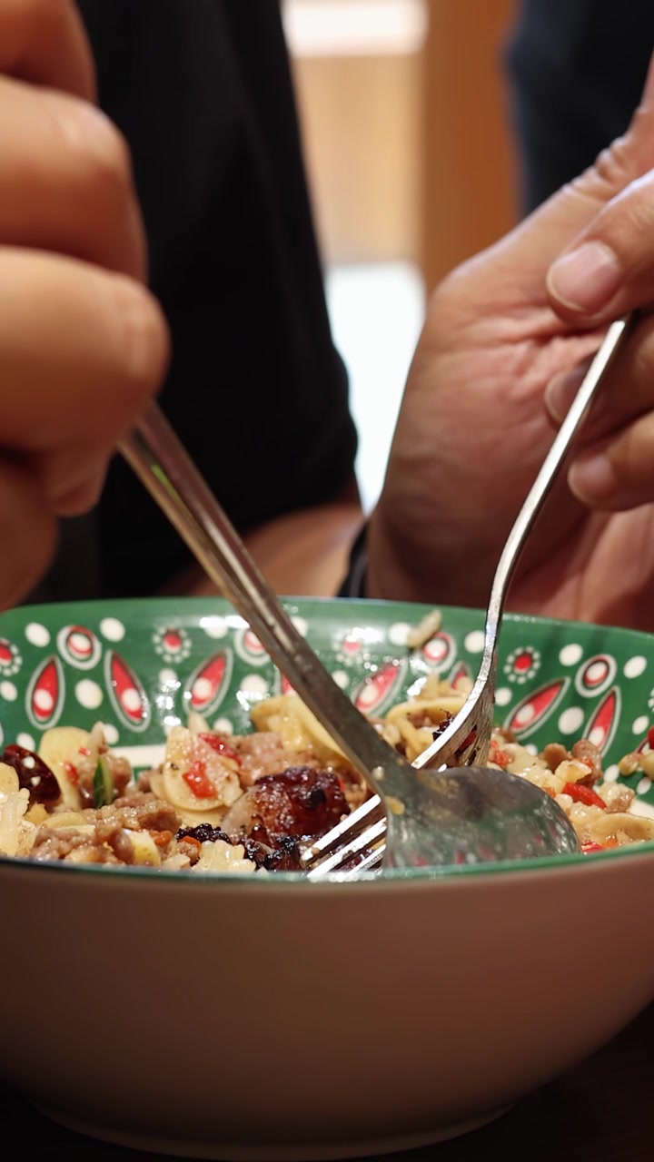 A person uses a spoon and fork to mix a colorful Thai rice dish in a patterned bowl under warm, natural indoor lighting with close-up framing