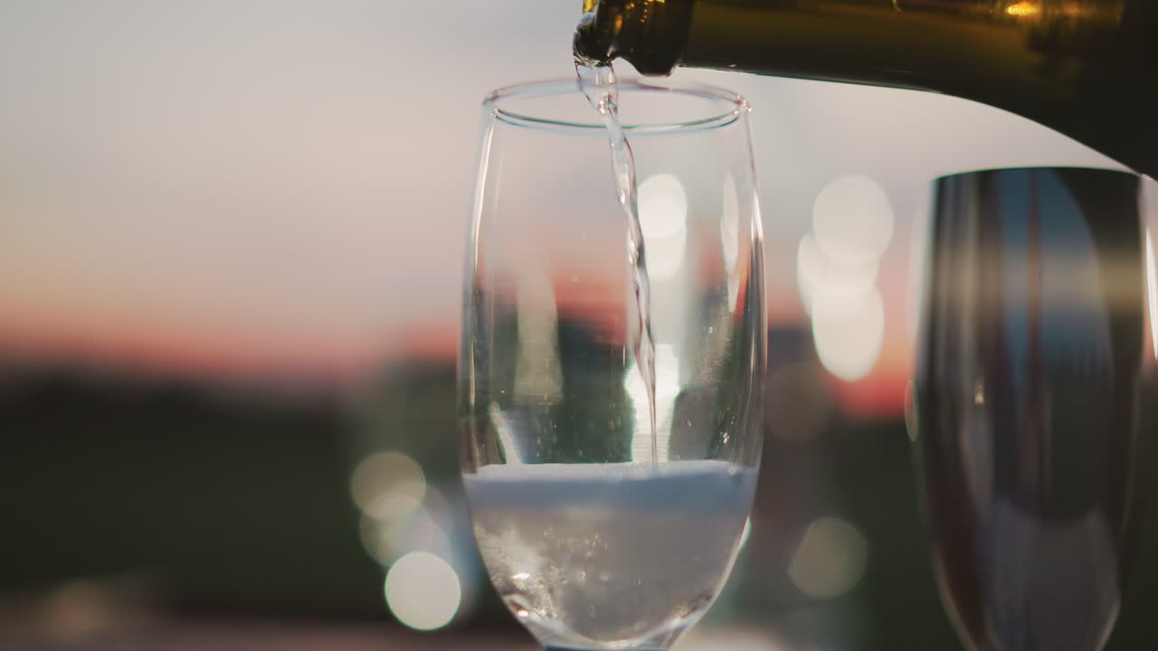close up of wine being poured into glass under sunset sky with sparkling bokeh lights in background capturing elegant pour motion and shimmering reflections against warm amber horizon glow