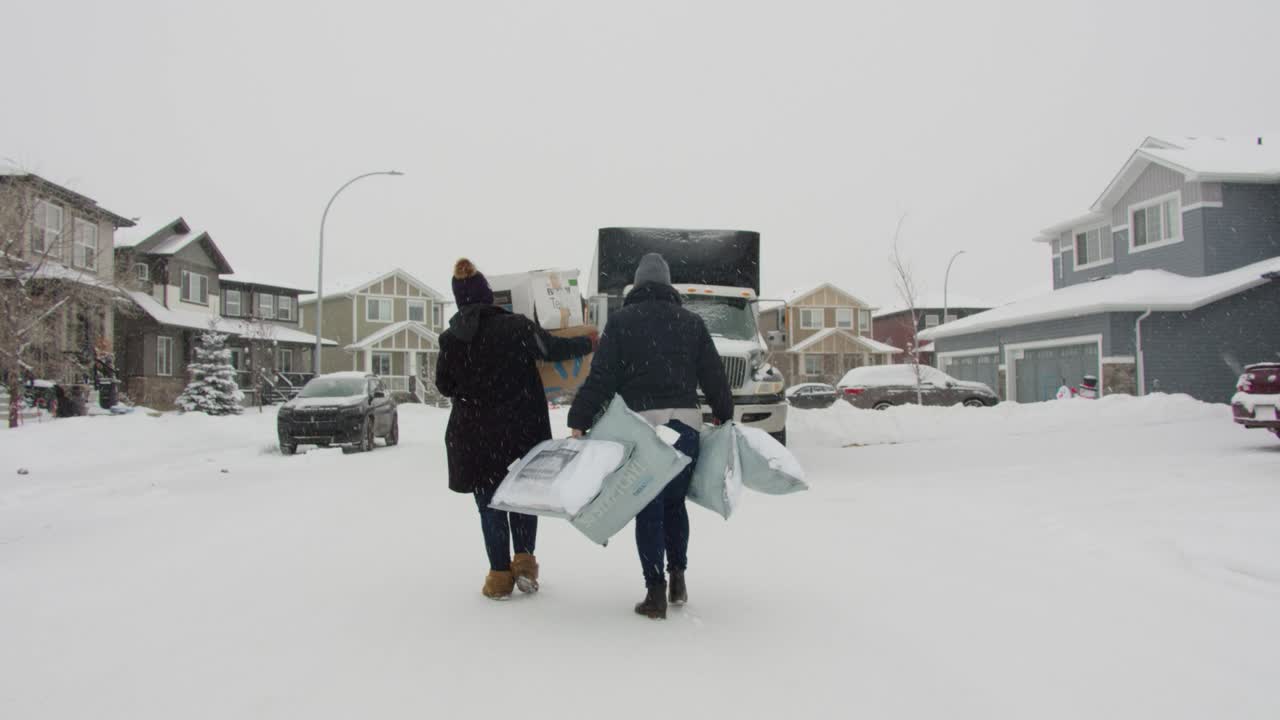 Slow motion shot two women walking away on snowy street in Canadian suburb carrying supplies, snow falling