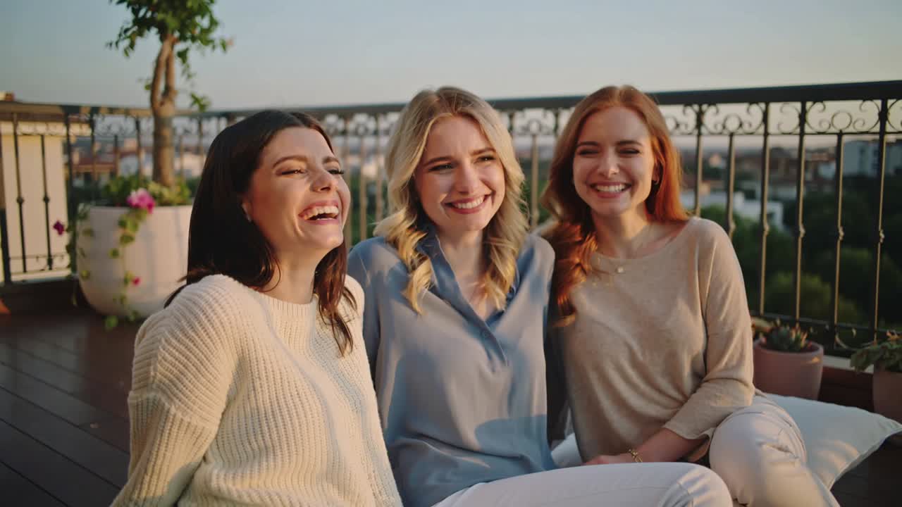 Tre amici che ridono, vestiti in color crema, blu e beige, appoggiati a una terrazza sul tetto, condividono uno scherzo vicino alla ringhiera.
