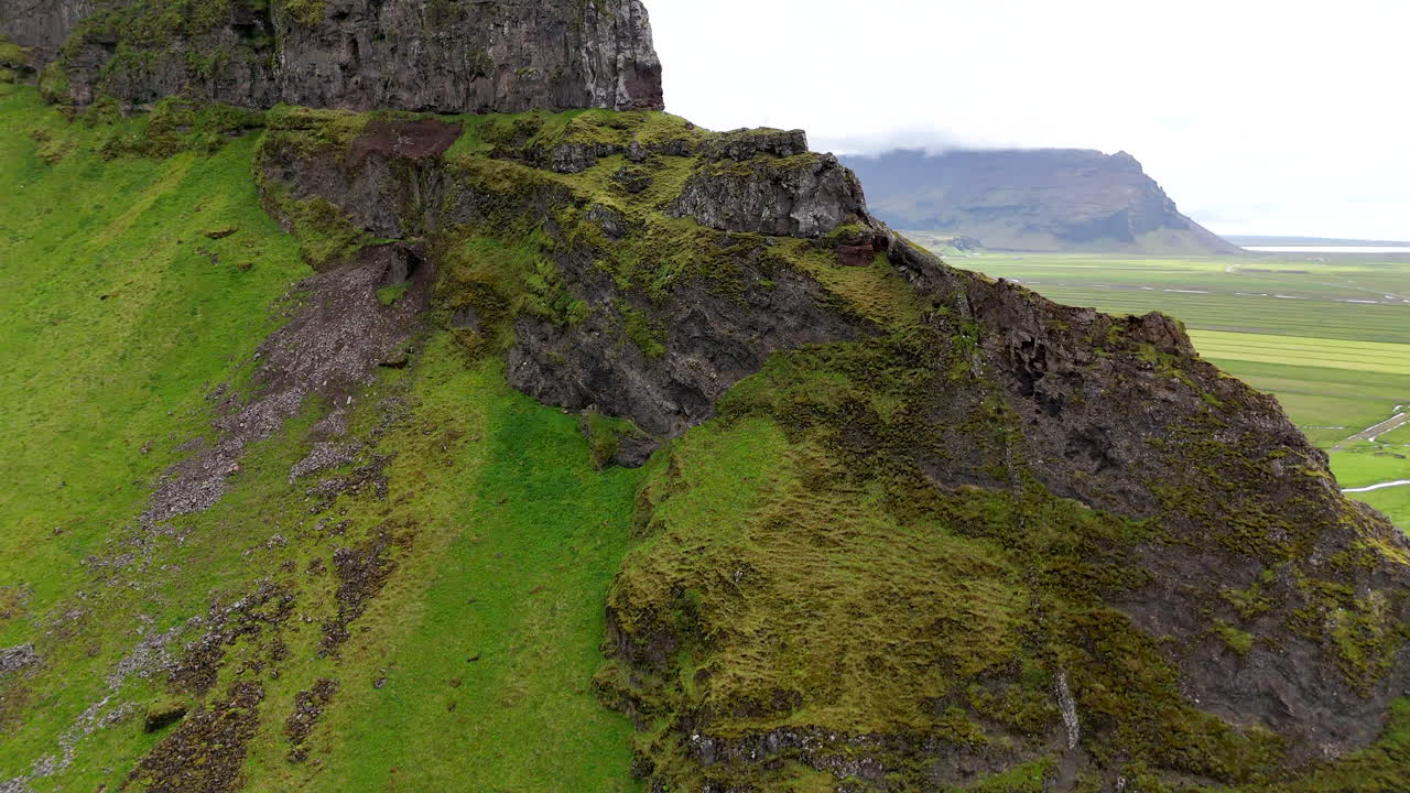 Aerial view of vibrant green fields stretching below steep moss-covered cliffs in Suðurland, Iceland, with winding streams and dramatic shadows shaping the vast volcanic landscape