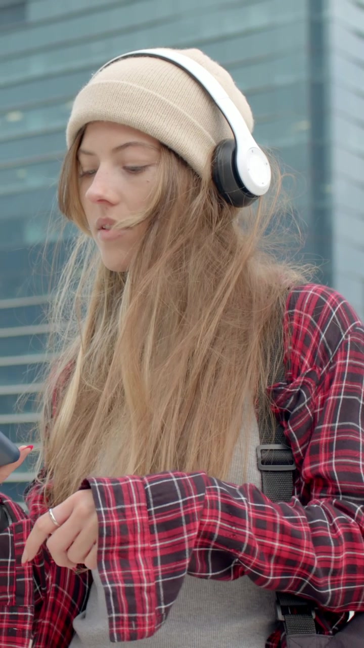 Video verticale: Donna con berretto che si sistema le cuffie dopo una folata di vento, controllando il telefono vicino a una parete di vetro.