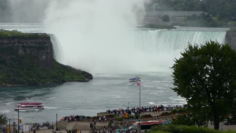 Tourists-boats-at-Niagara-Falls,-next-to-powerful-water-flowing-down-the-waterfall-creating-steam,-on-a-moody-day