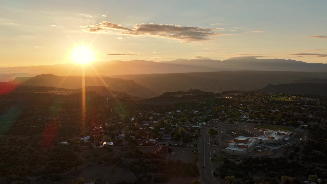 Timelapse 4K con drone di un'alba vibrante che inonda un tranquillo quartiere di White Rock, Nuovo Messico. Raggi di sole dorati e bagliori dell'obiettivo illuminano i vasti canyon del deserto d'alta quota, la catena montuosa del Sangre de Cristo