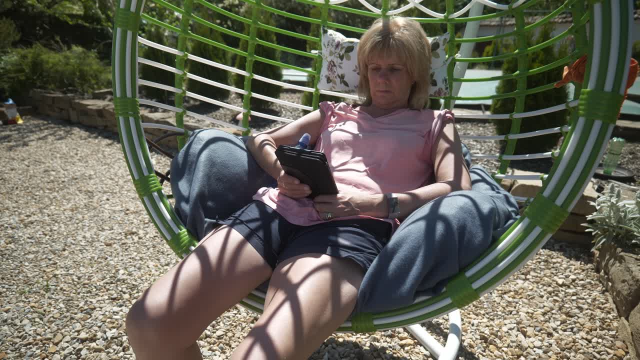 mujer rubia en pantalones cortos lee la novela de libros electrónicos relajándose en un cómodo columpio de hamaca en el jardín en un día caluroso y soleado