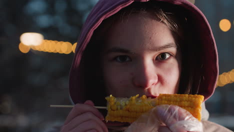 lady eating corn on stick wrapped in white plastic with bokeh lights in blurred background, steam rising from fresh corn in outdoor setting, enjoying snack on street