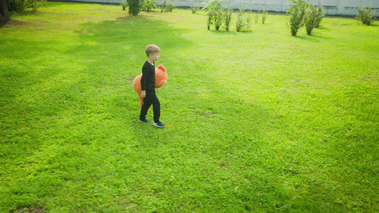 Lonely child wearing black outfit walking quietly and thoughtfully across sunlit green grassy field holding large orange teddy bear, surrounded by scattered bushes, trees, and white fence