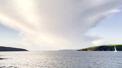 Lonely-sailing-boat-on-leaving-safety-of-the-harbour-in-Kinsale,-Ireland-with-dramatic-cloud-over-blue-sky