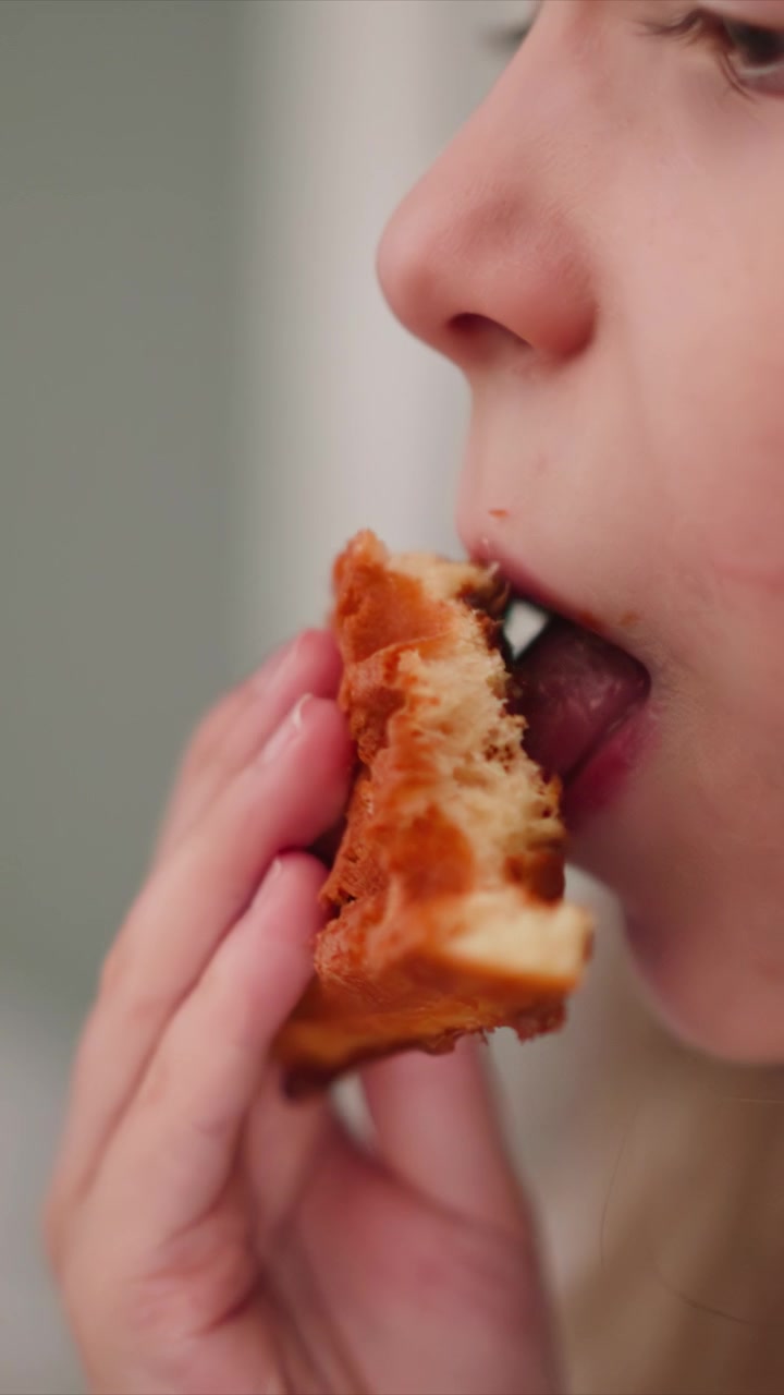Close up of young woman eating waffle. Hand holding golden brown food with soft lighting and natural expression. Indoor setting with blurred background emphasizing subject profile