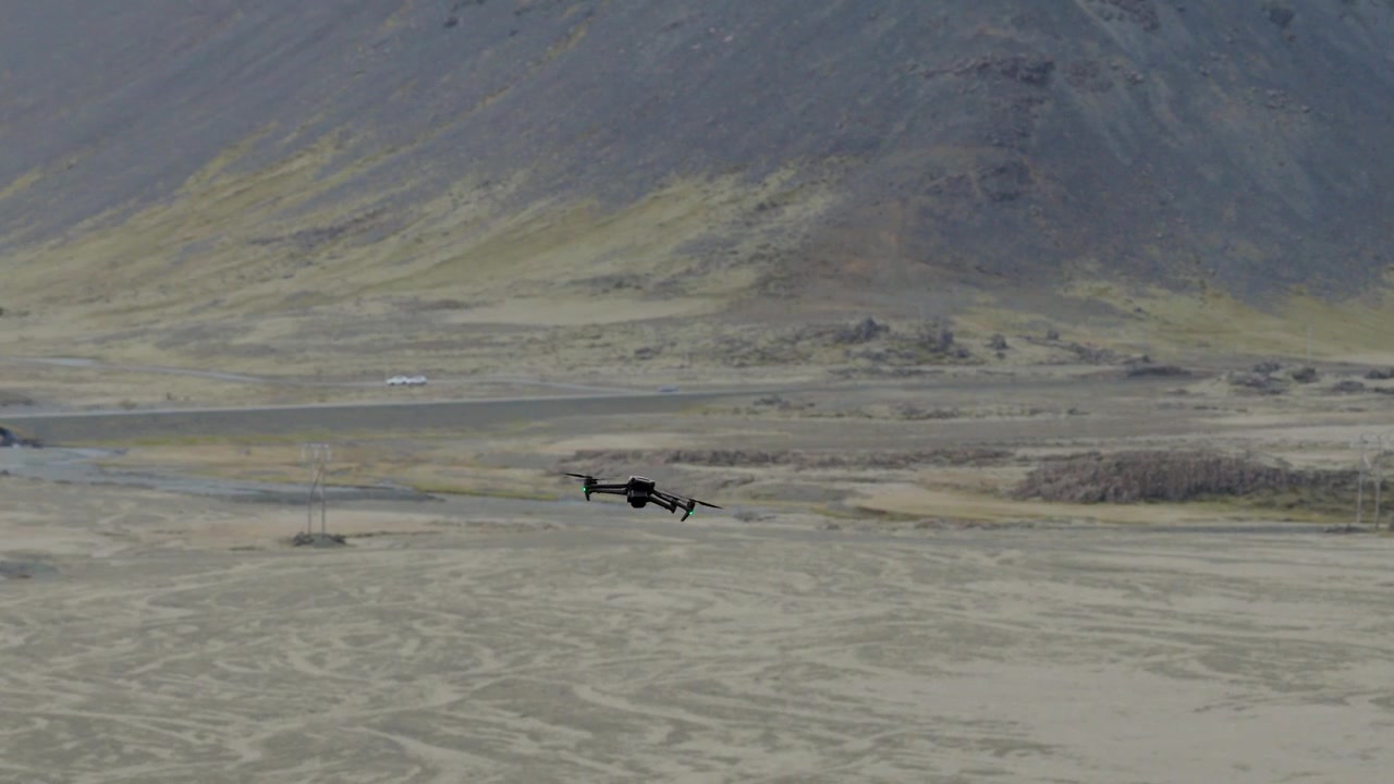Camera drone flies low above pale glacial outwash with gravel deposits and braided sediment channels beneath steep volcanic mountain slopes, Iceland, drone tracking.