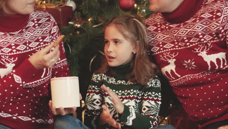 Happy-Family-In-Christmas-Sweaters-Sitting-Together-In-Front-Of-Christmas-Tree-At-Home-While-Mother-Feeding-Little-Girl-With-A-Cookie