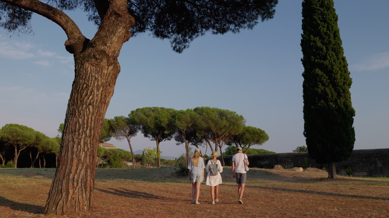 Una famiglia che passeggia nel Parco dell'Appia Antica a Roma sotto i pini con l'antico acquedotto e una calda luce serale.
