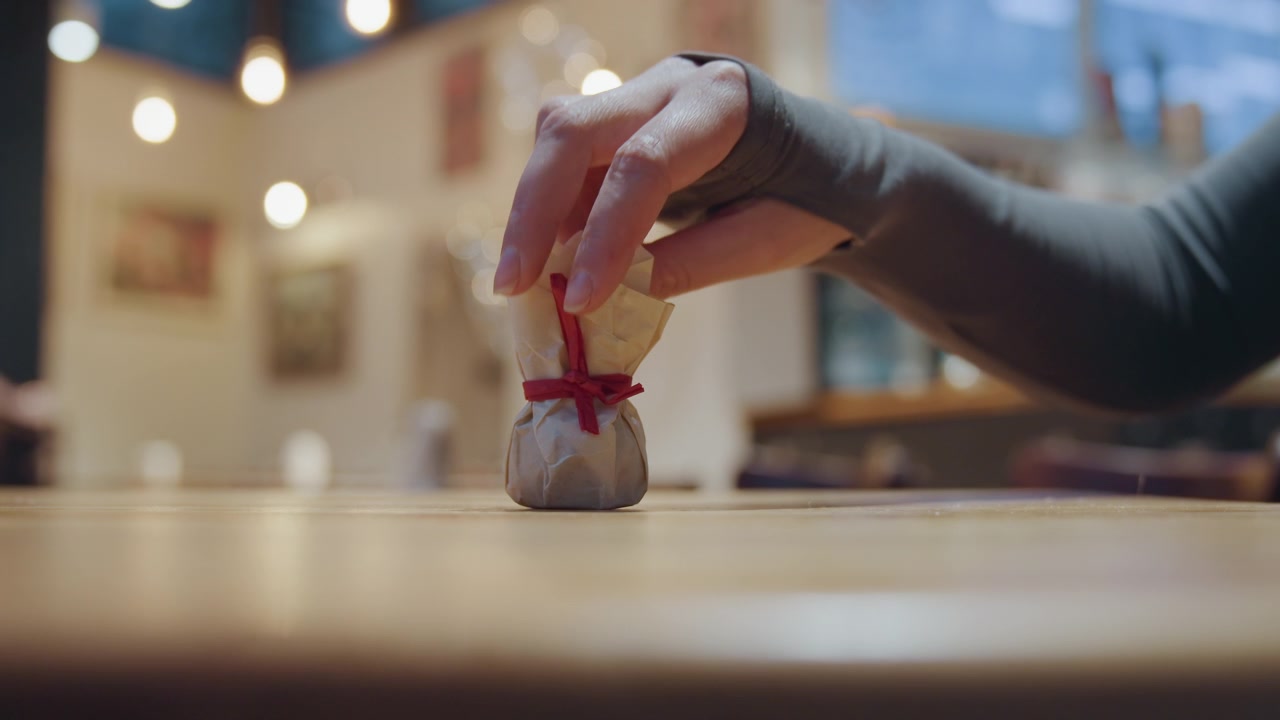 Caucasian hand picking pastry from closed cafe table, small ribbonwrapped sweet on wooden surface, delicate grasp and reaching fingers, empty dining room with cordon and warm lights, intimate closeup
