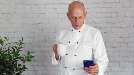 A-mature-senior-head-chef-is-taking-a-break-from-the-kitchen-and-texting-on-his-phone-while-drinking-coffee