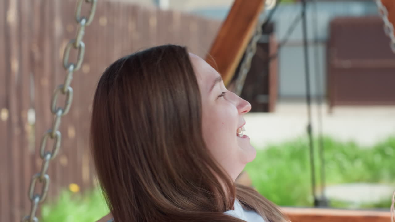Close up of student girl sitting peacefully on wooden swing in outdoor backyard, sunlight lights her face as she gazes ahead, with shopping bag beside her and background of fence and greenery