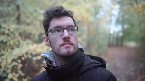 joven europeo con gafas y barba de pie en un bosque de otoño mixto, mirando a la distancia