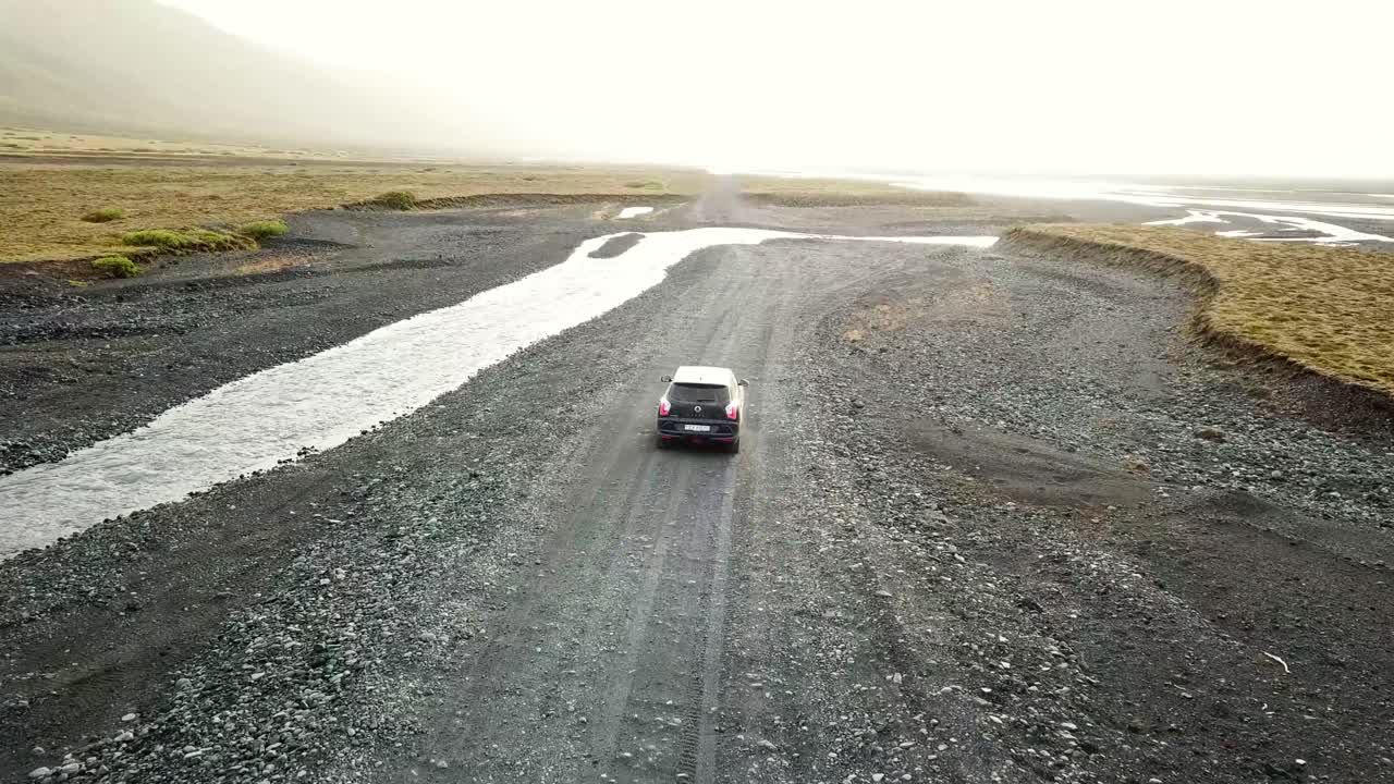 A car navigates a remote gravel road in Þórsmörk, Iceland, surrounded by rugged terrain, small streams, and golden light from the sunrise