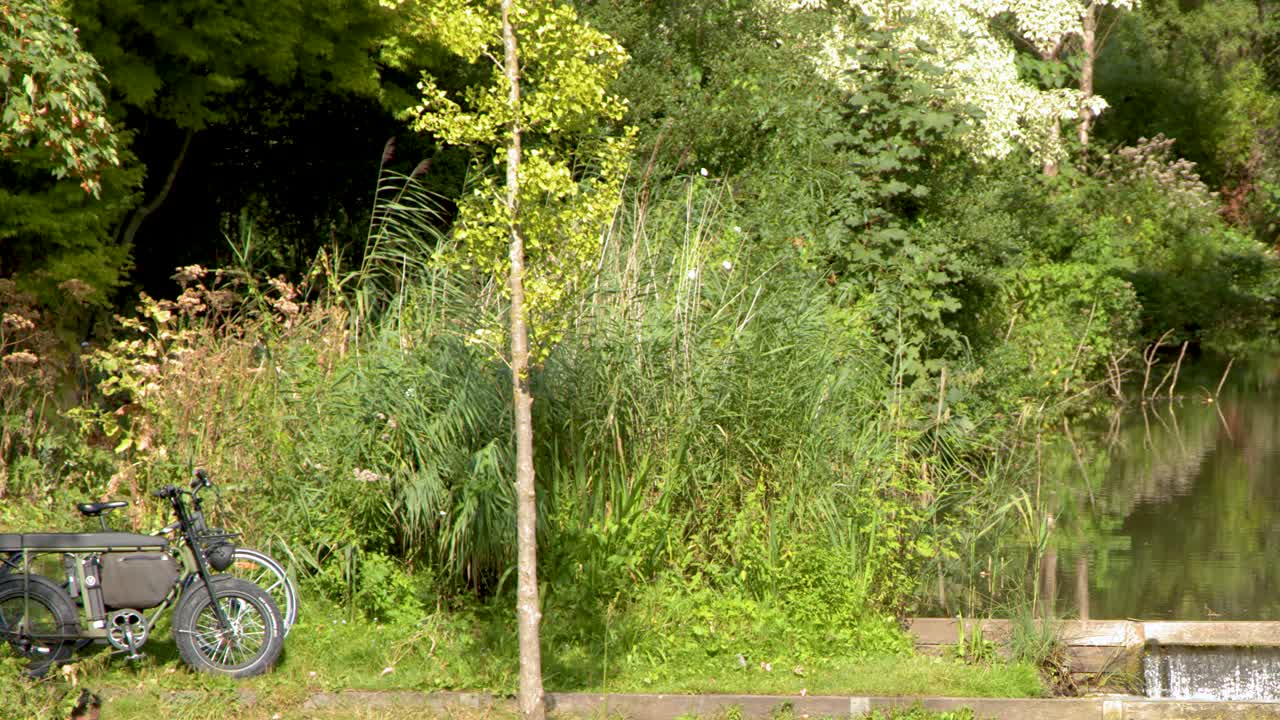Electric bicycle rides past canal and dam, lush greenery, steady daylight, smooth horizontal camera pan