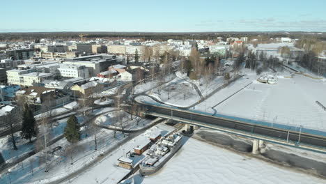 Antena,-Seguimiento,-Toma-De-Drones,-Panorámica-Desde-El-Paisaje-Urbano,-Sobre-El-Tráfico-En-El-Puente-Suvantosilta,-Sobre-El-Río-Pielisjoki,-En-Un-Día-Soleado-De-Invierno,-En-Joensuu,-Karelia-Del-Norte,-Finlandia