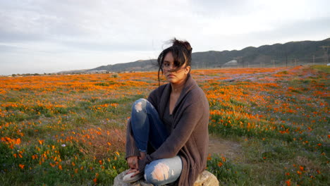 a beautiful young woman crying and looking heartbroken and depressed sitting alone in a rural field of wild flowers with wind blowing her hair at sunset slow motion