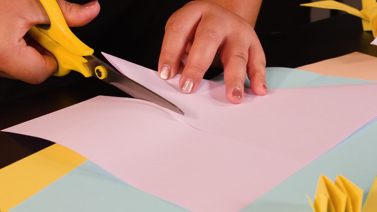 Primo piano di mani che usano forbici gialle per tagliare carta rosa su una scrivania colorata