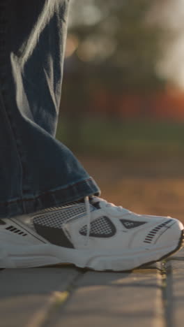 un primer plano de una persona con vaqueros y zapatos blancos, caminando lentamente por un camino pavimentado en un parque. la luz cálida y suave y el fondo borroso