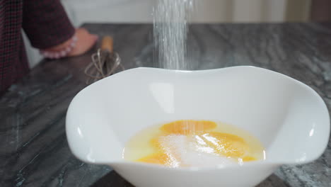 close-up of sugar being poured into eggs in a white bowl on a dark countertop, a person hand rests on the table, adorned with a bracelet, while a whisk lies nearby