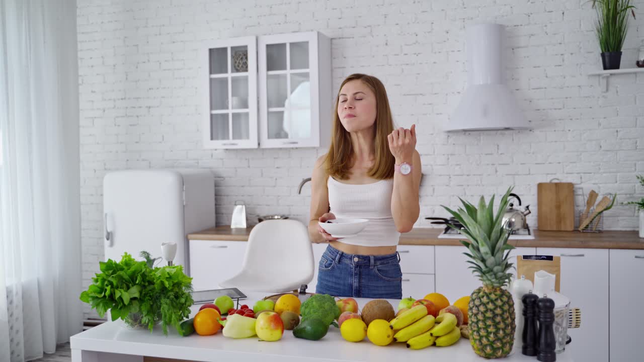 Young woman in the modern kitchen. Beautiful female is eating while standing at the table full of organic vitamins. Healthy lifestyle. Dieting concept.