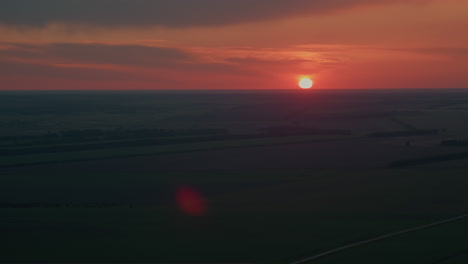 bright orange sun nears horizon casting warm glow across layered evening sky filled with soft clouds as distant farmland stretches beneath creating calm tranquil mood