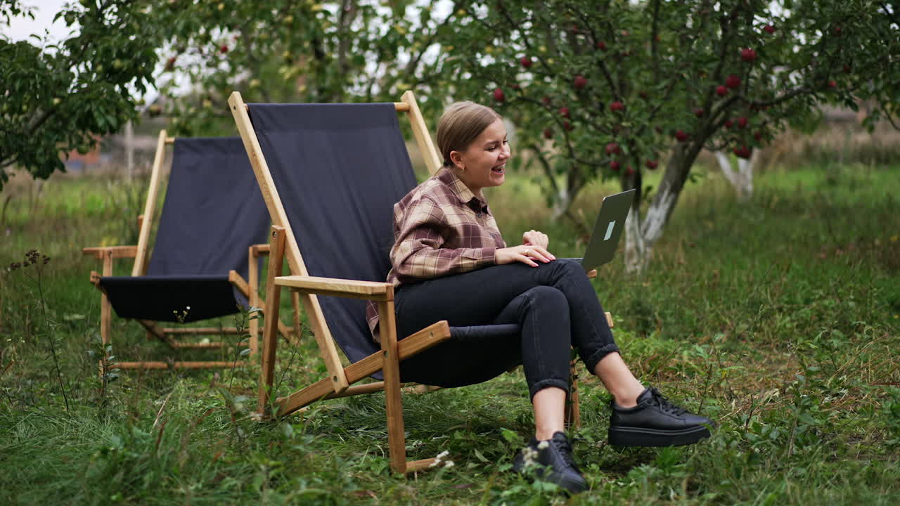 Caucasian woman watching video on laptop sitting outdoors. Lady is smiling happily and then looks astonished. Nature backdrop.