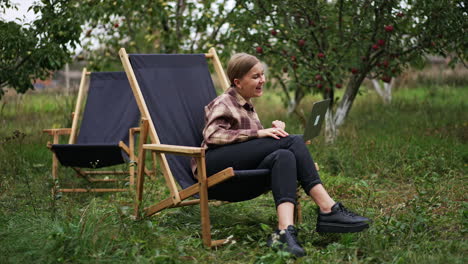 caucasian woman watching video on laptop sitting outdoors. lady is smiling happily and then looks astonished. nature backdrop.