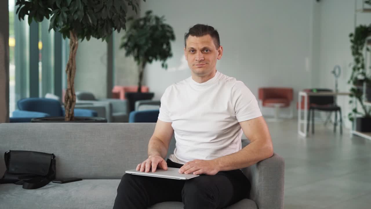 ⁇  티셔츠 를 입은 청년 이 비즈니스 센터 를 배경 으로 노트북 에서 일 을 마치고 카메라 를 쳐다보고 미소 짓고 있는 클로즈업 초상화