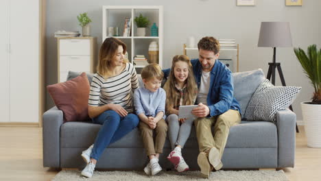 Familia-Alegre-Con-Dos-Niños-Sentados-En-El-Sofá,-Sonriendo-Mientras-Ve-Algo-En-La-Tableta