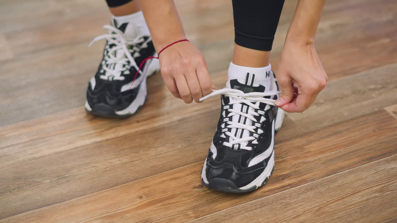 Leg view of girl wearing black leggings bending down tying shoelaces of white and black sneakers, red bracelet visible on wrist, detail focused on hands adjusting lace properly on wooden floor