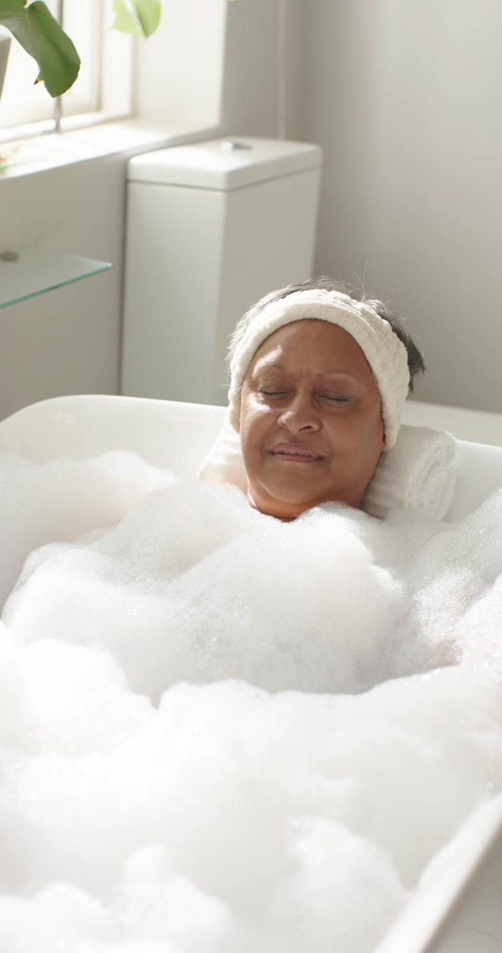Vertical video: Mature woman reclining in bathtub at home with foam, head on towel, seeking rest