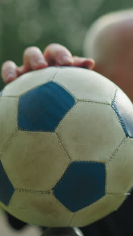 una vista borrosa de un anciano con una camiseta negra moviendo suavemente una pelota de fútbol con su mano, con una vista borrosa de una carretera pavimentada y un campo cubierto de hierba