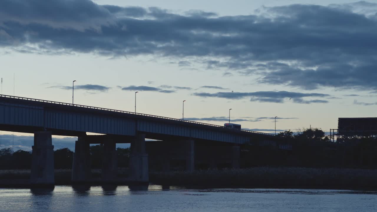 A semi-truck drives across a long bridge over calm river waters during twilight, under a moody blue sky