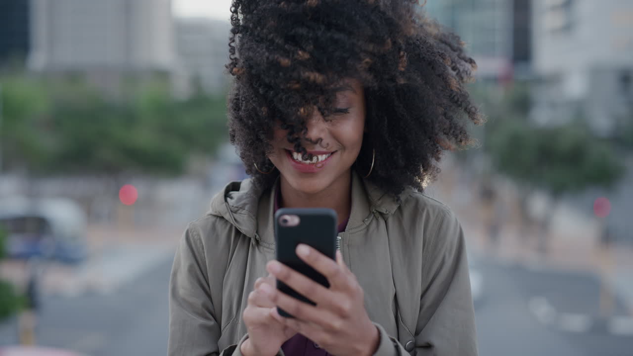 ritratto di una felice donna afroamericana che usa lo smartphone per inviare messaggi in città sorridendo godendosi la navigazione dei messaggi online sul cellulare vento che soffia acconciatura afro