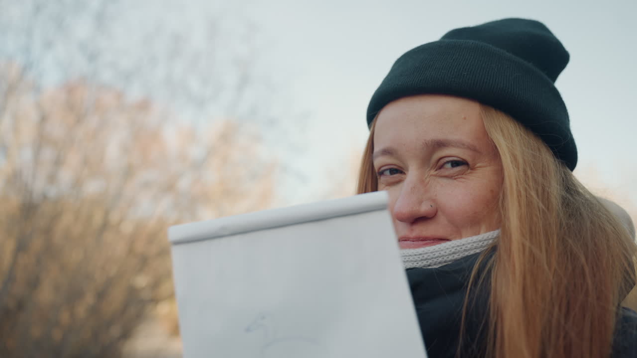 Ritratto di una donna bianca dai capelli rossi che sorride all'aperto in un parco, rivelando uno schizzo su un blocco da disegno. L'artista, con berretto, sciarpa e giacca, mostra un primo piano del viso e della mano che tiene un foglio con un semplice disegno di anatra, in un'atmosfera calda.