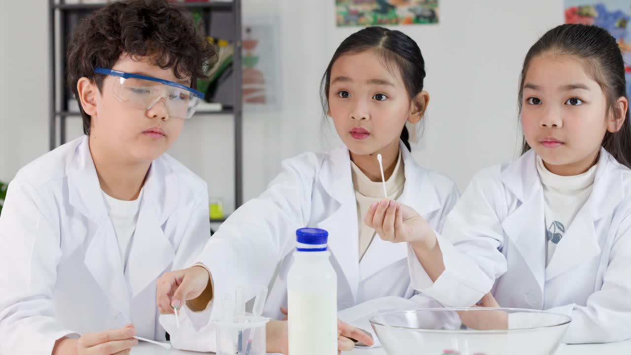 3人の若いアジア人学生が、白衣と保護メガネを着用して化学実験を行っている。