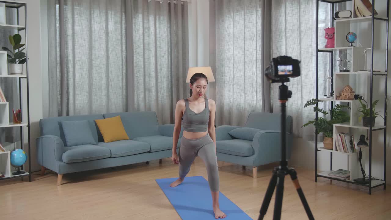 스포츠 옷을 입은 젊은 아시아 트레이너 여성이 카메라에 대화하고 집에서 운동을 가르치는 것을 녹음하는 동안 높은 지 자세로 요가를합니다.