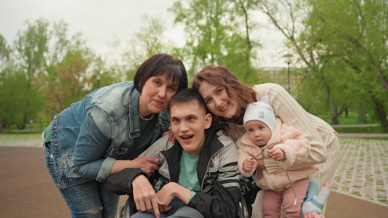 White Family Smiling Together On Park Path, Mother And Sister Leaning Over Son In Wheelchair Holding Baby Candid Joyful Outing, Playful Interaction, Supportive Care And Bright Spring Greenery