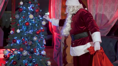 authentic santa putting gifts under christmas tree. red boxes with presents at new year's night. new year tradition. santa near beautiful christmas tree