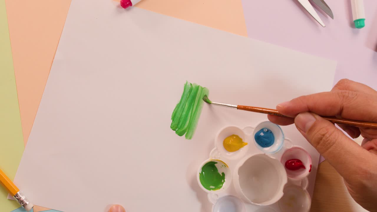 Artist hand paints green strokes on white paper using brush, palette, and primary color paints