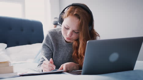 female college student wearing headphones works on bed in shared house with laptop and mobile phone