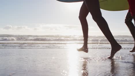 Unterer-Abschnitt-Eines-Afroamerikanischen-Paares,-Das-Mit-Surfbrettern-Am-Strand-Läuft