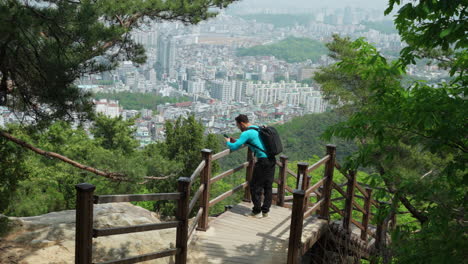 Mann-Wanderer,-Der-Beim-Wandern-Auf-Dem-Gwanaksan-Bergweg-Sein-Telefon-Benutzt,-Mit-Blick-Von-Oben-Auf-Das-Skyline-Panorama-Von-Seoul-Im-Hintergrund-–-Filmisches-Einschieben-Des-Gimbals