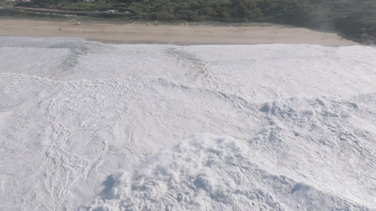 Aerial drone shot of big waves coming into shore on a day with giant waves in Nazaré, Portugal, Europe. Flying towards beach and people watching. Big wave surfing town