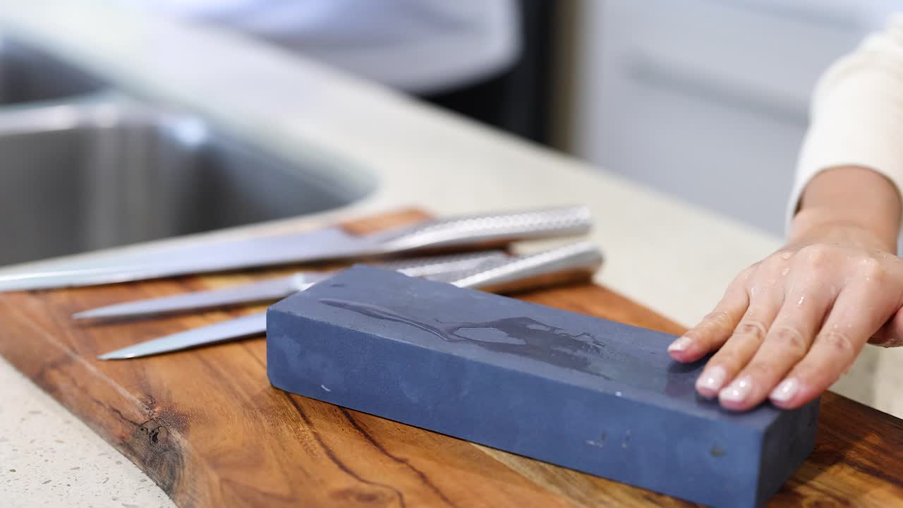 A person sharpens knives using a whetstone on a wooden board in a well-lit kitchen